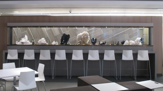A view of the dramatic long, narrow exhibit case filled with GIA Museum pieces. The case is a feature wall in the student commons area filled with tables and chairs.